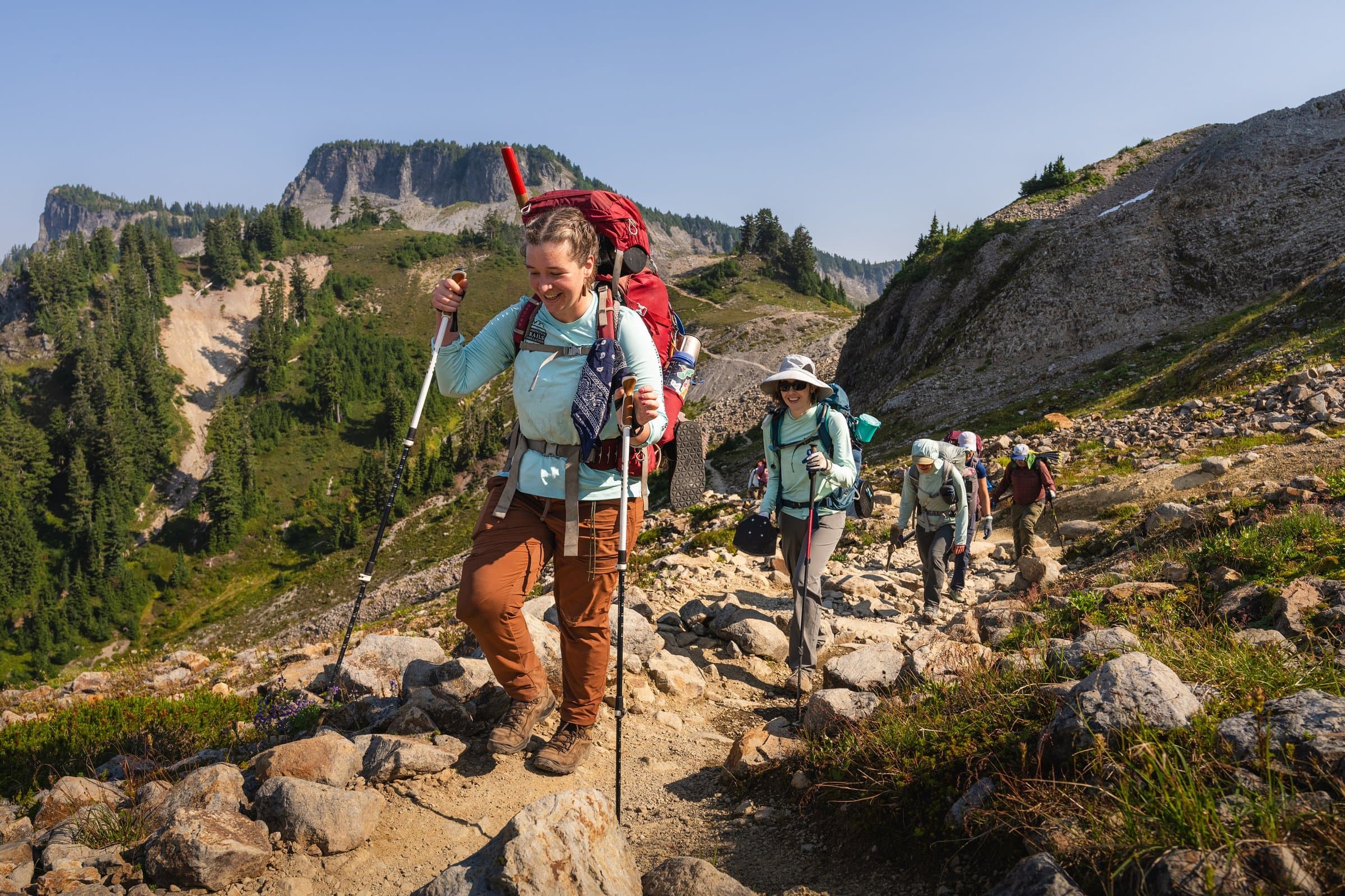 Five backpackers hiking up a trail lined by rocks with mountain views in the background.